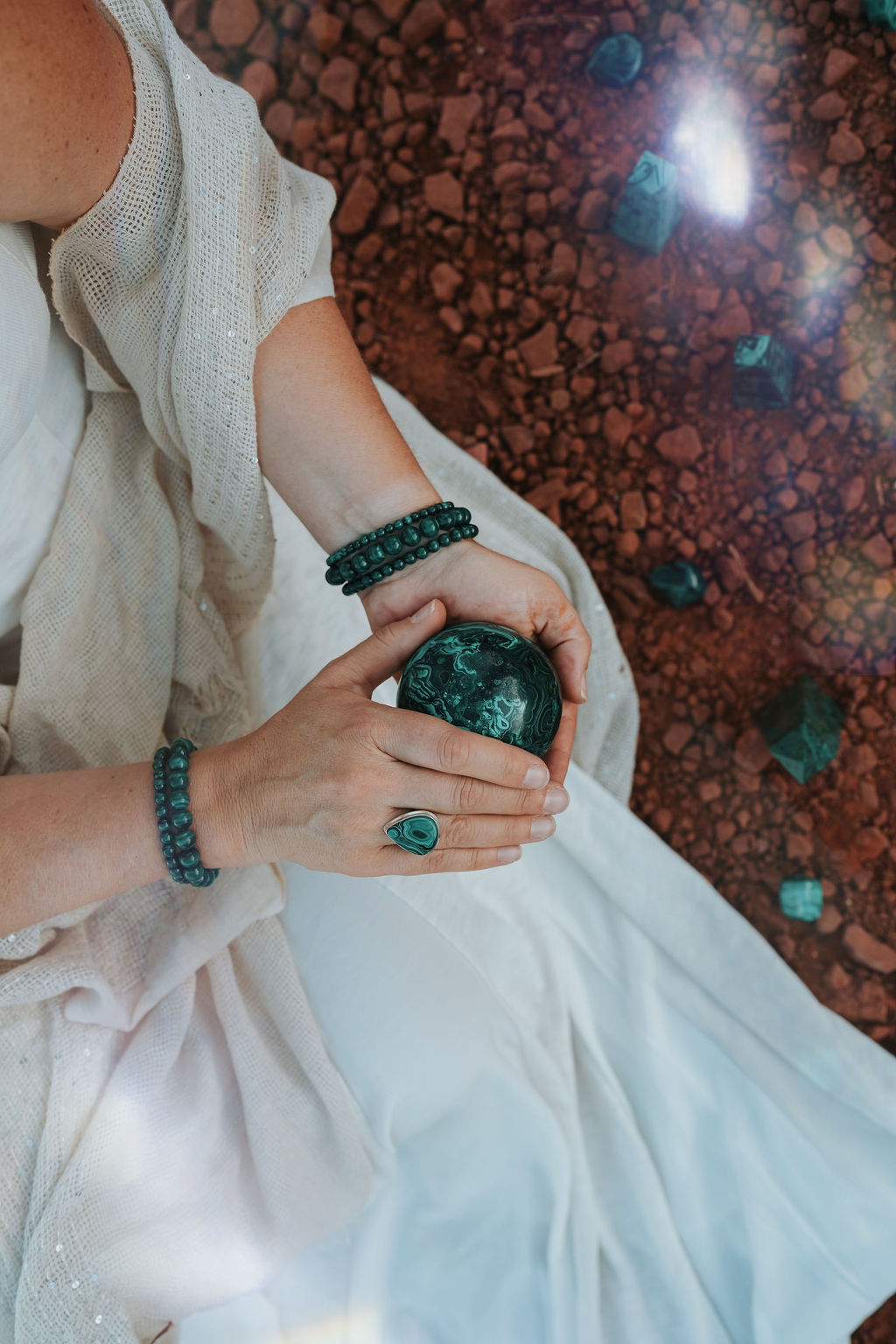 Hands holding a malachite crystal sphere with matching malachite ring and bead bracelets on a white dress, Sedona red rock backdrop.